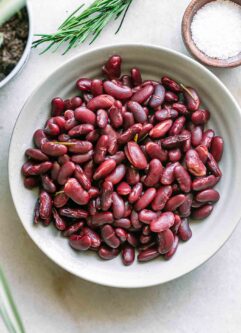 a bowl of cooked kidney beans on a white table with herbs and bowls of seasonings
