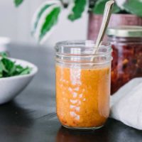 a mason jar with tomato salad dressing on a wooden table with a bowl of arugula and green plant