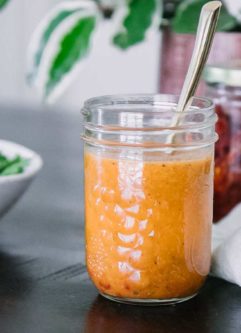 a mason jar with tomato salad dressing on a wooden table with a bowl of arugula and green plant