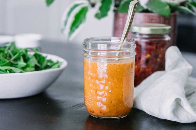 a mason jar with tomato salad dressing on a wooden table with a bowl of arugula and green plant