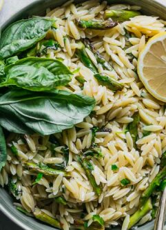 a bowl of lemon asparagus orzo with fresh herbs on a white table with a lemon