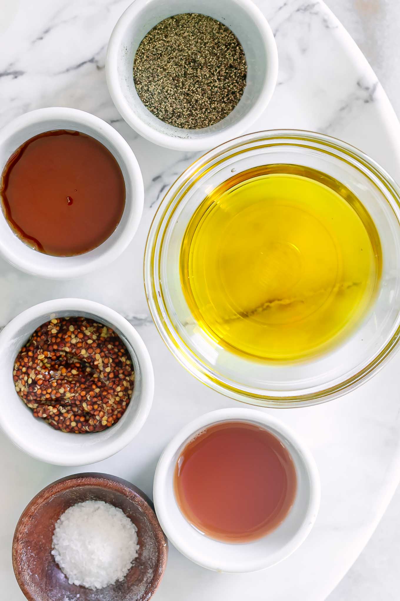 bowls of olive oil, dijon mustard, maple syrup, salt, pepper, and apple cider vinegar on a white table
