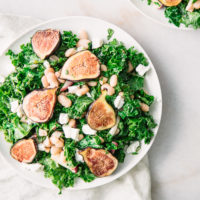 A green salad with kale, figs, white beans, and goat cheese on a white plate.
