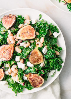 A green salad with kale, figs, white beans, and goat cheese on a white plate.