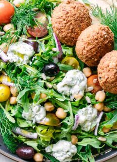 an Mediterranean-style salad with arugula, falafel, chickpea, tomatoes, onion, hummus, tzatziki, and sunflower seeds on a blue plate