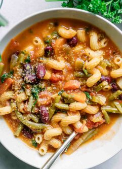a close up photo of minestrone soup with kidney beans and cavatappi pasta noodles with a gold fork