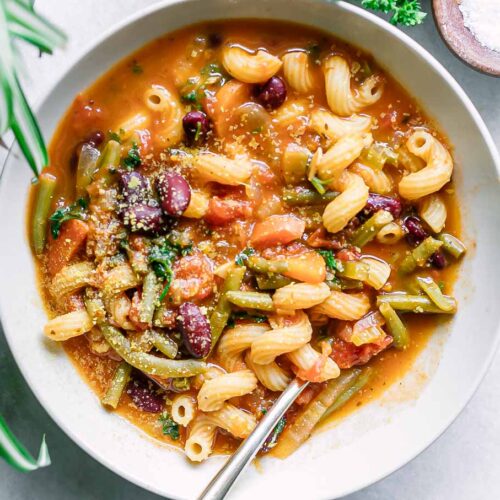 a white bowl of vegan minestrone soup on a white table with a gold fork