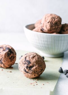 a chocolate brownie bite ball on a white table with a bowl of more snack balls in the background