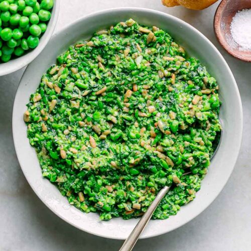 a bowl of green pea pesto on a white table with a bowl of fresh spring peas