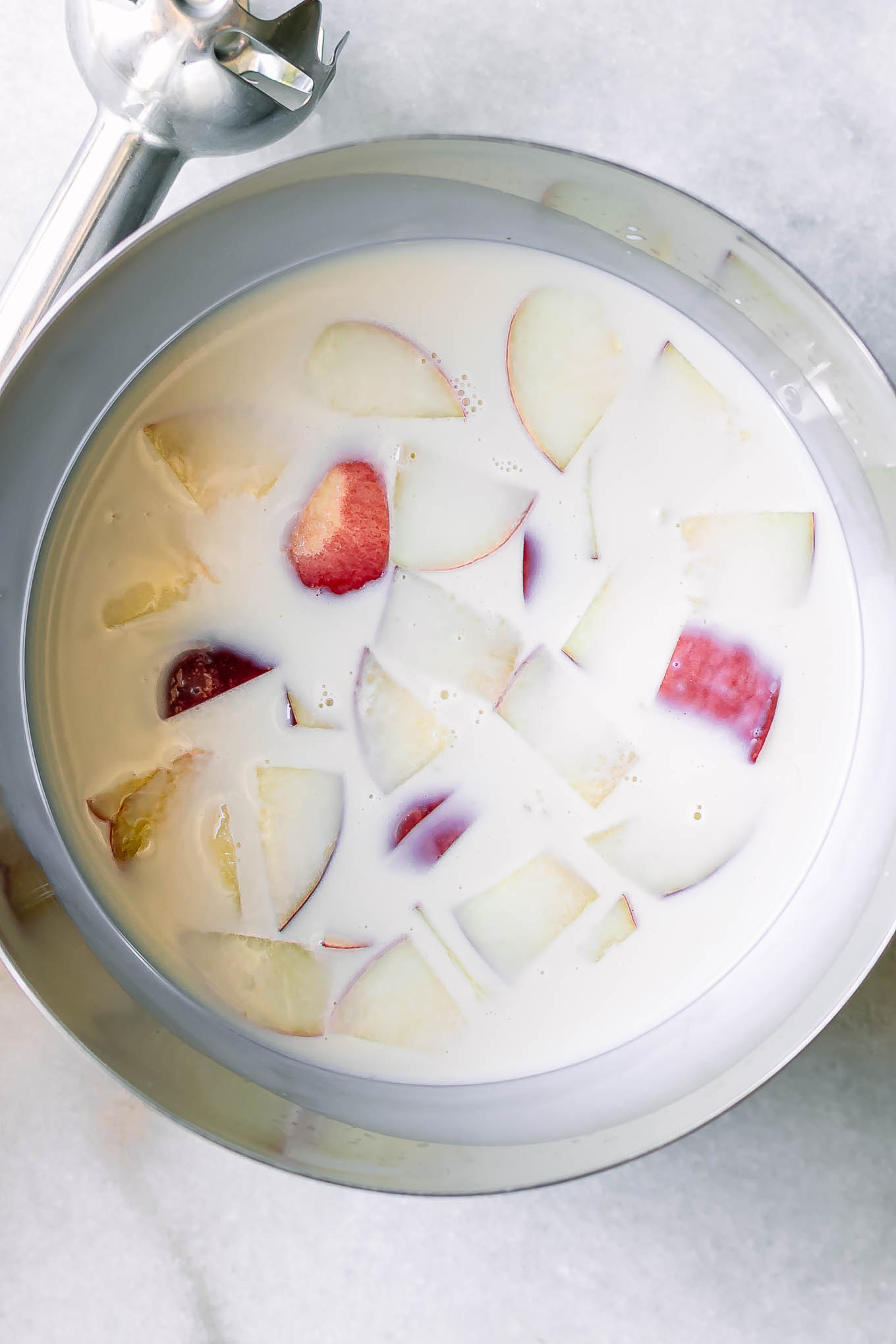 a large mixing bowl with plant-based milk and sliced peaches on a table with an immersion blender