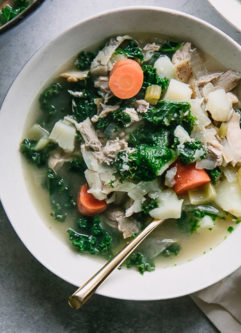 a bowl of chicken soup with potatoes, kale, and carrots on a blue table with a half a lemon