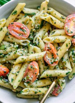 a white bowl with pesto penne pasta and cherry tomatoes on a white table with a gold fork