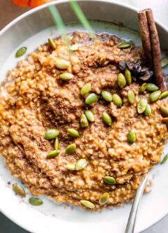a bowl of pumpkin oatmeal with pumpkin seeds as a garnish and a cinnamon stick on a white table with a mini pumpkin