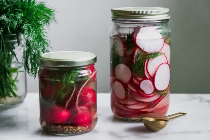 two jars of sliced and whole pickled radishes on a white table