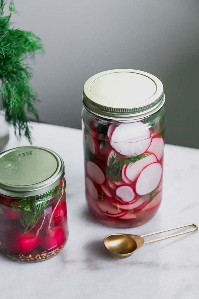 two jars of sliced and whole pickled radishes on a white table with fresh dill