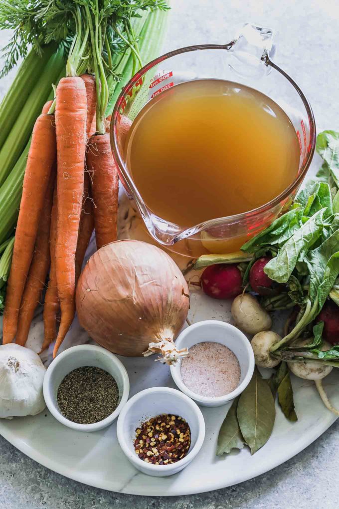 radishes with greens, carrots, celery, onion, garlic, and bowls of spices and vegetable broth on a white table for soup