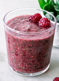 a glass with a spinach raspberry smoothie on a white table with fresh strawberries and a bowl of fresh spinach