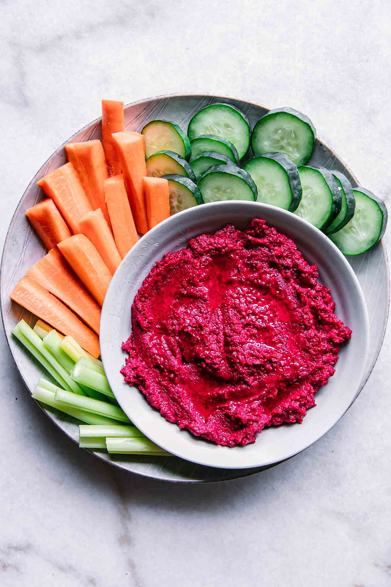 a bowl of red hummus with beets on a plate with cut cucumbers, carrots, and celery on a white table