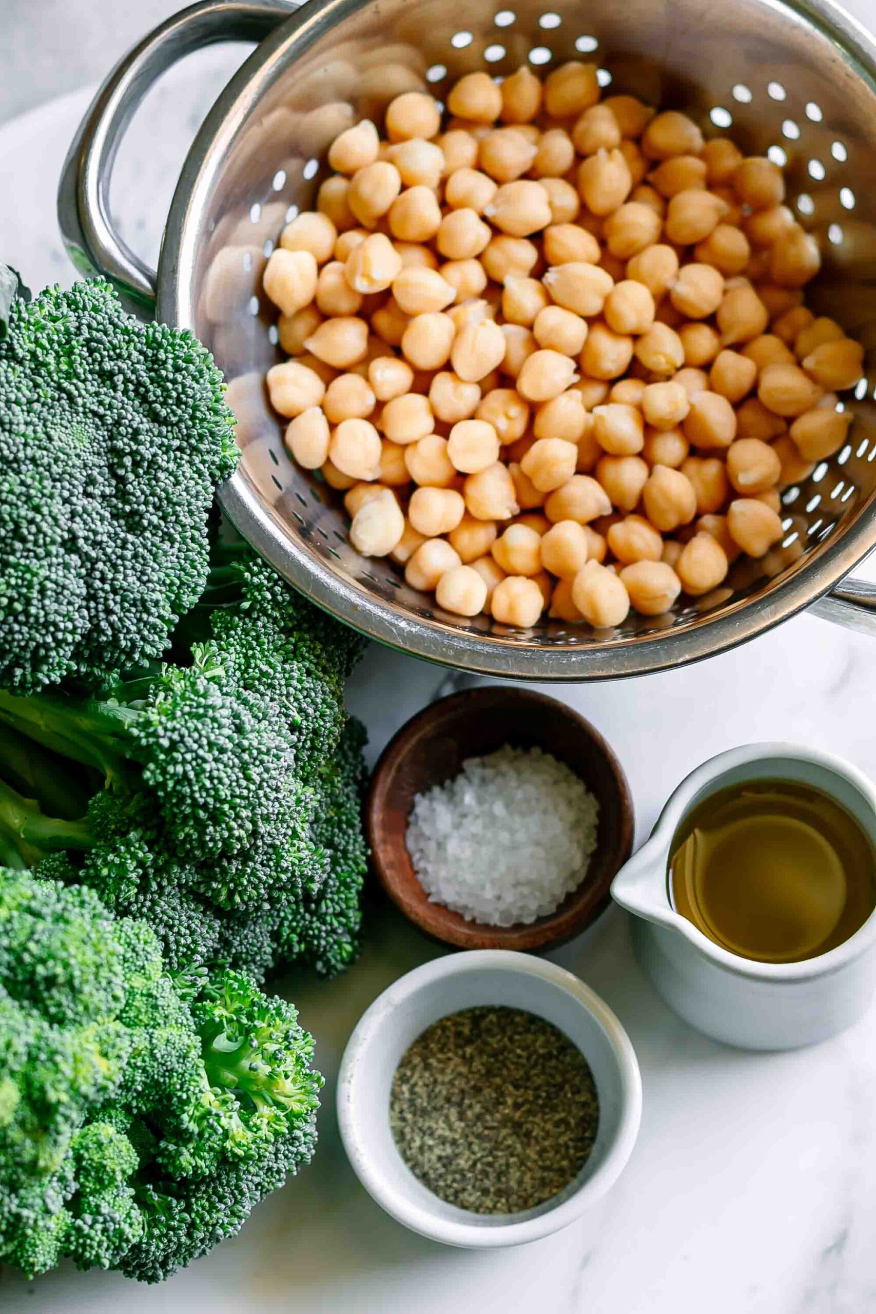 a bunch of broccoli, a colander with chickpeas, and bowls of olive oil, salt, and pepper