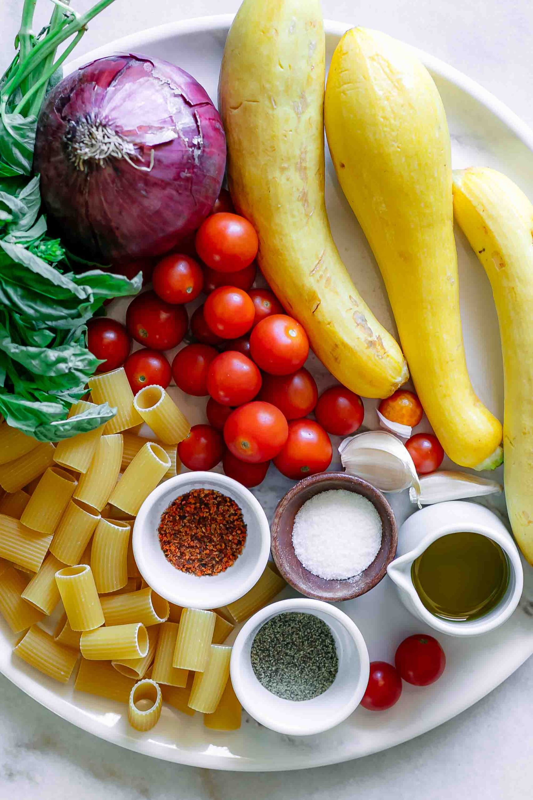 summer squash, cherry tomatoes, onions, basil, garlic, olive oil, spices, and dry pasta on a white table