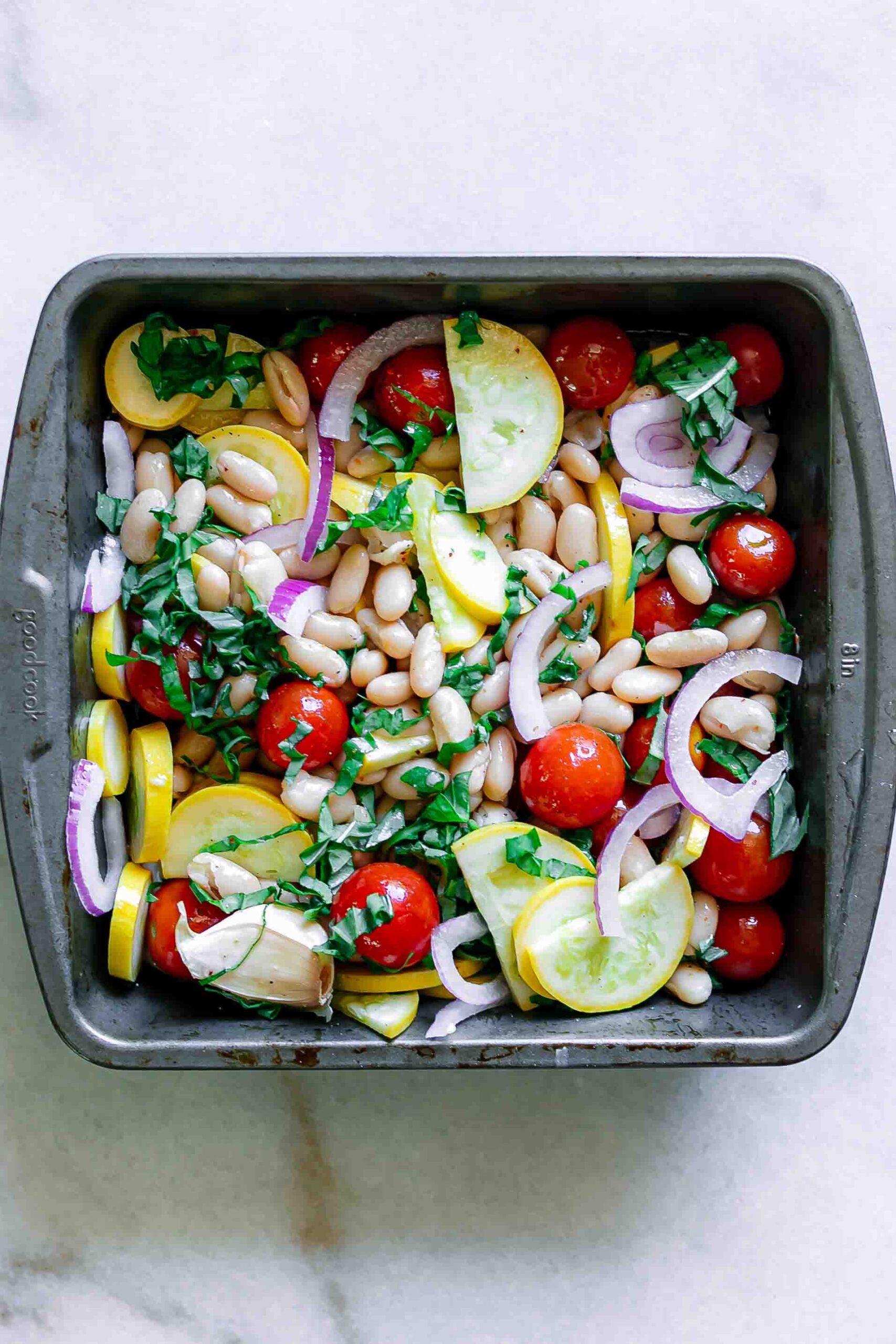 a squash roasting pan with squash, cherry tomatoes, red onion, basil, and spices on a white table before roasting