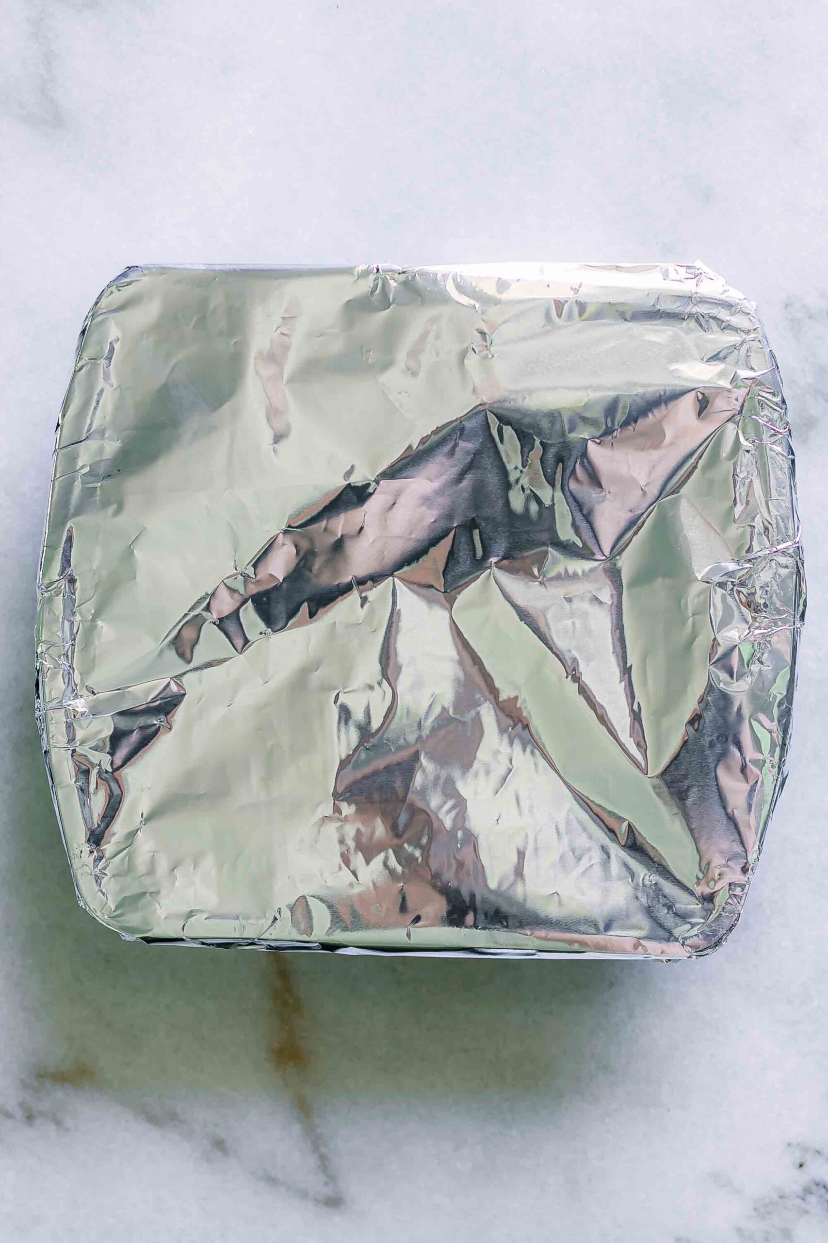 a roasting pan with vegetables covered with aluminum foil on a white table