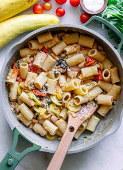 a pan with rigotoni pasta in a sauce of creamy white beans, yellow summer squash, cherry tomatoes, fresh basil, and garlic on a white table