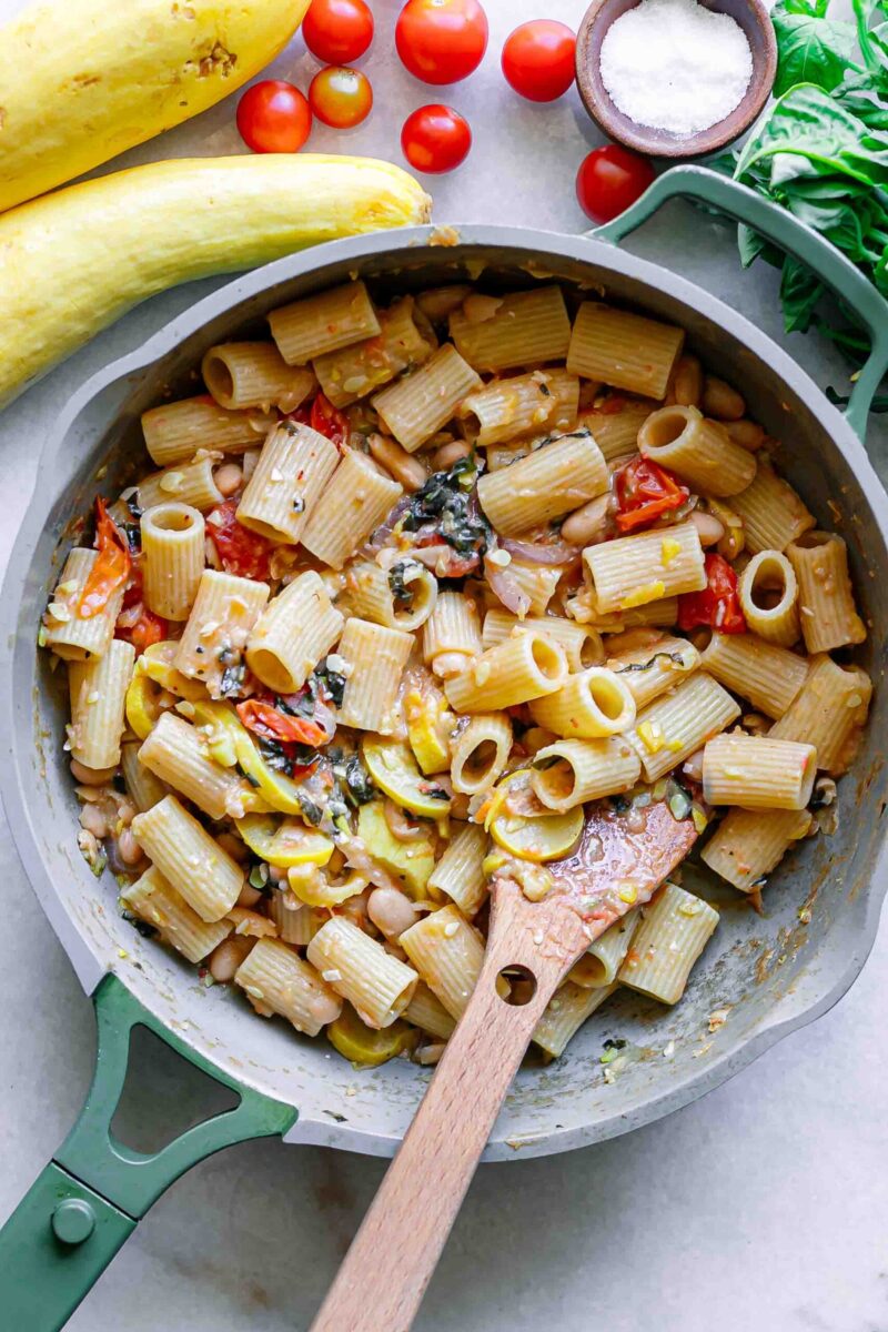 a pan with rigotoni pasta in a sauce of creamy white beans, yellow summer squash, cherry tomatoes, fresh basil, and garlic on a white table
