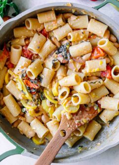pasta in a creamy white bean sauce with roasted squash, cherry tomatoes, onions, and basil on a white table with a wooden spoon