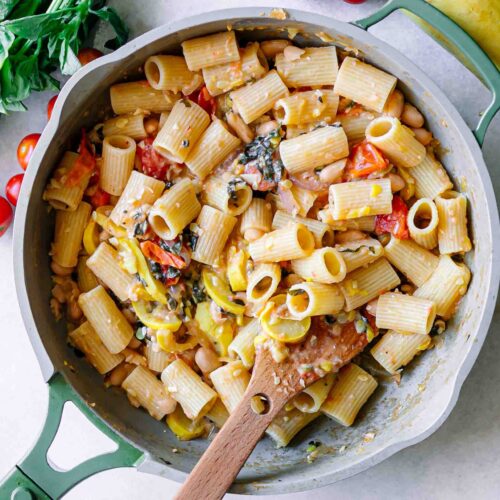 pasta in a creamy white bean sauce with roasted squash, cherry tomatoes, onions, and basil on a white table with a wooden spoon