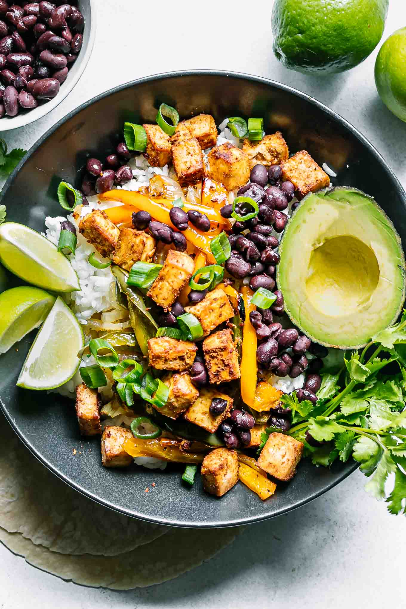 a tofu, vegetable, and rice fajita bowl with black beans, limes, and avocado on a white table