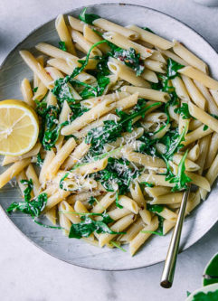 a plate of penne pasta with spinach and a slice of lemon on a white table with a bowl of parmesan cheese