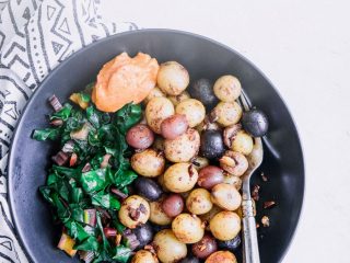 A black bowl with breakfast potatoes, cooked greens, and garlic aioli.