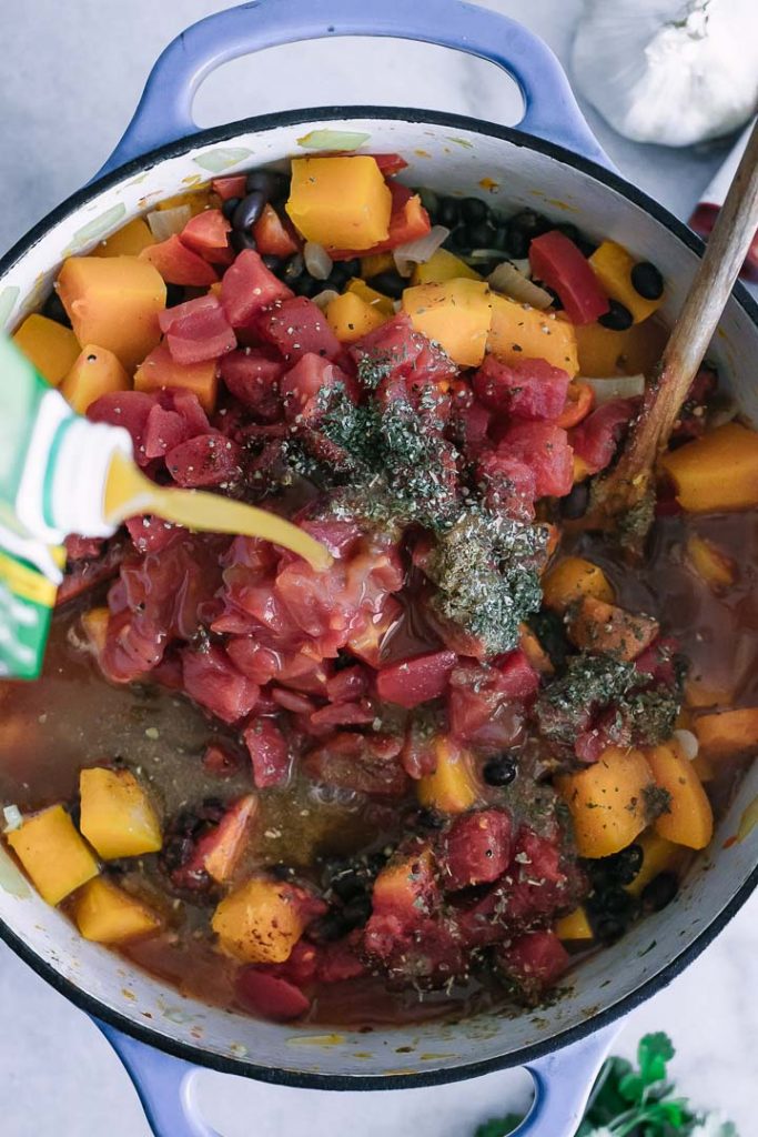 vegetable broth pouring into a large soup pot with butternut squash, tomatoes, and black beans