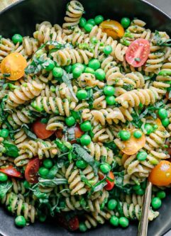 a bowl of pesto pasta salad with green peas, tomatoes, and basil on a white table with a gold fork
