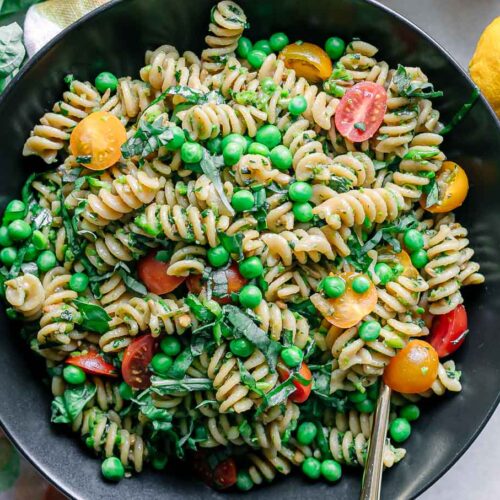 a bowl of pesto pasta salad with green peas, tomatoes, and basil on a white table with a gold fork