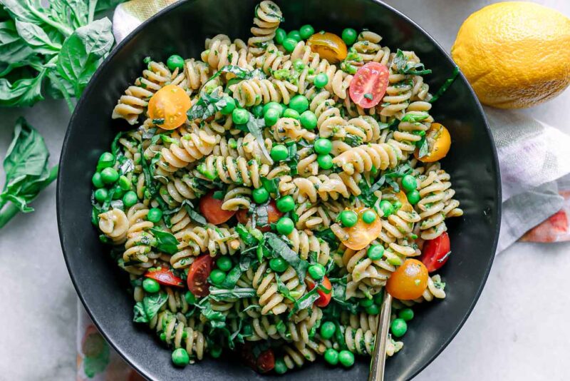 a bowl of pesto pasta salad with green peas, tomatoes, and basil on a white table with a gold fork