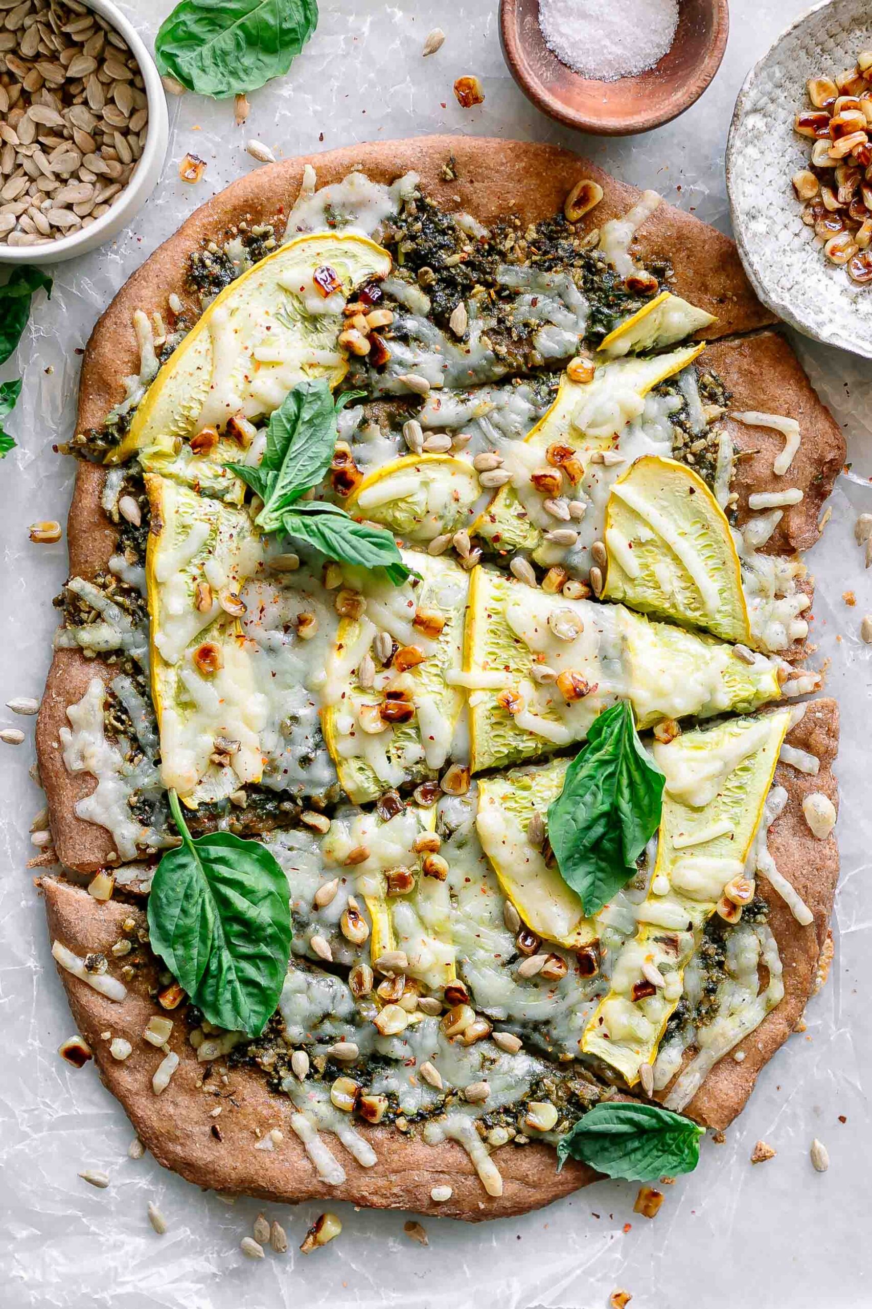 a pizza with yellow squash, pesto, corn, and basil on a white table