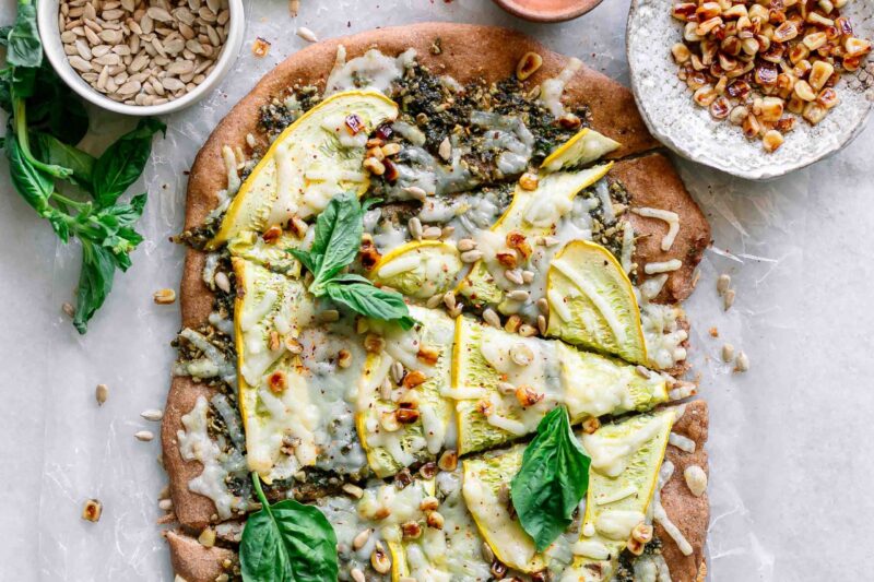 a pizza with sunflower seed pesto, sliced yellow summer squash, vegan cheese, a sprinkle of corn, and fresh basil on top