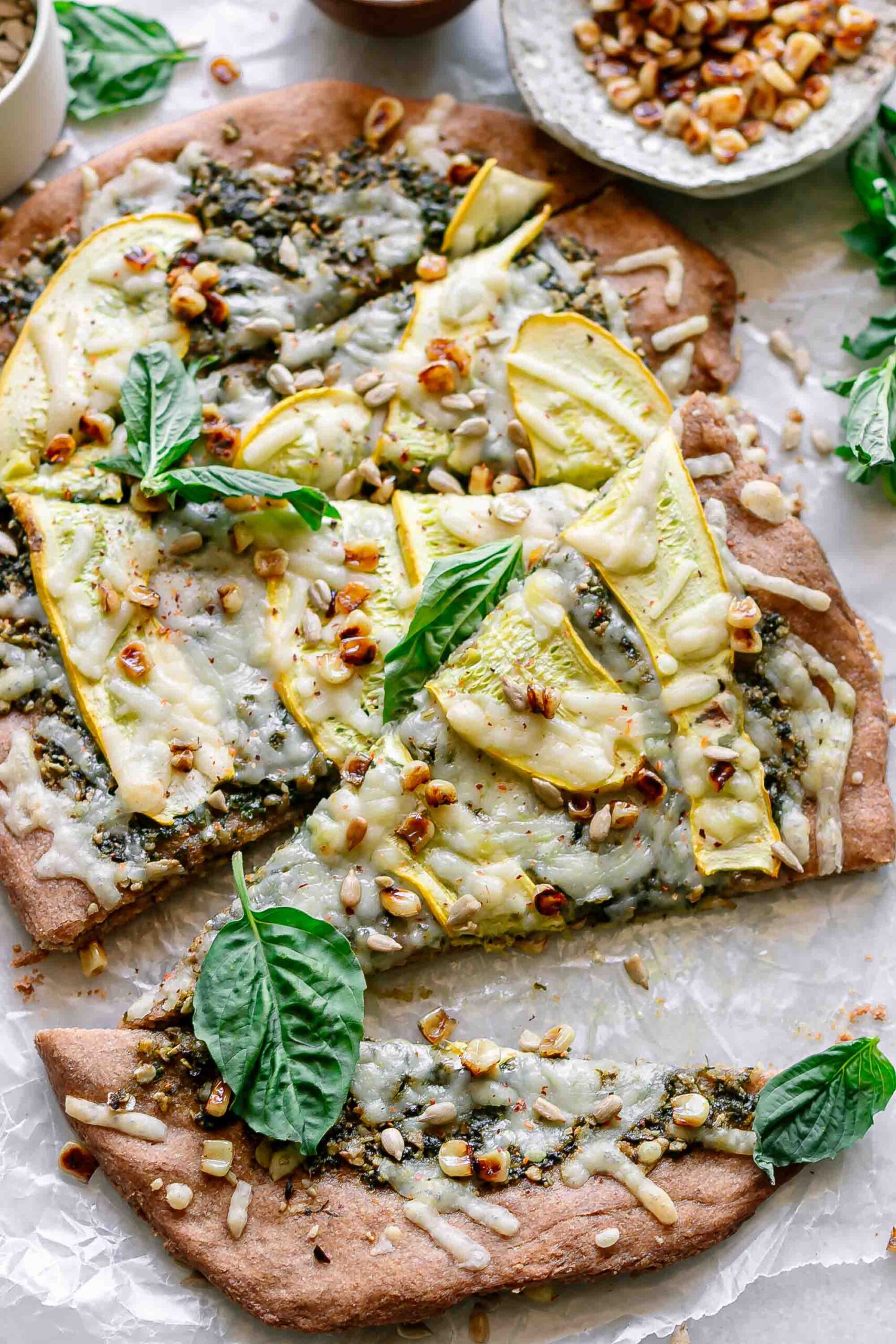 a pesto pizza with squash, corn, and basil cut into pieces on a white table