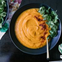 A bowl of orange soup made with sweet potatoes, carrots, and spicy chipotle with cilantro on top on a dark table