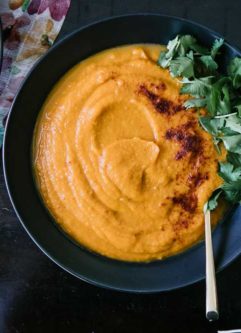 A bowl of orange soup made with sweet potatoes, carrots, and spicy chipotle with cilantro on top on a dark table