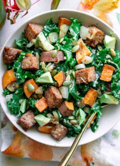 a white bowl with kale and quinoa salad with sweet potatoes and tempeh with avocado on a white table