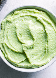 a bowl of vegan avocado crema sauce on a white table with an avocado