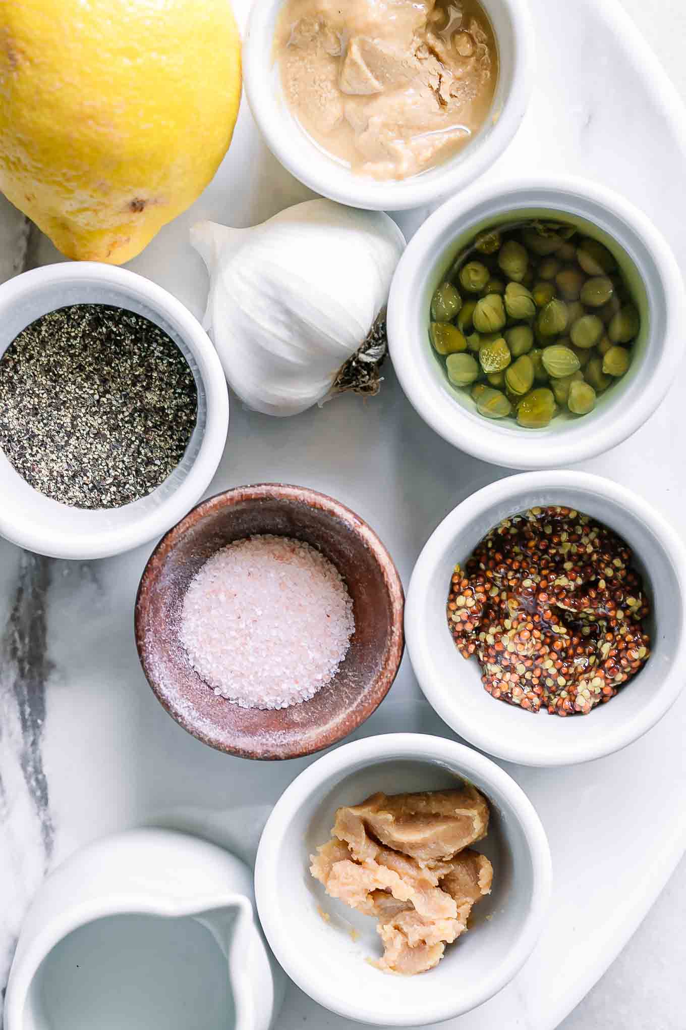 bowls of tahini, garlic, lemon, capers, mustard, white miso, water, and pepper on a white table