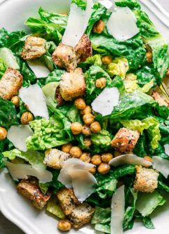 a white plate with vegan caesar salad with croutons, chickpeas, and shaved vegan parmesan cheese on a white table