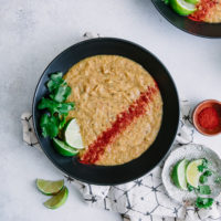 An orange corn and potato plant-based soup topped with cilantro and lime on a white table with a white and black napkin.