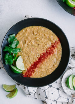 An orange corn and potato plant-based soup topped with cilantro and lime on a white table with a white and black napkin.
