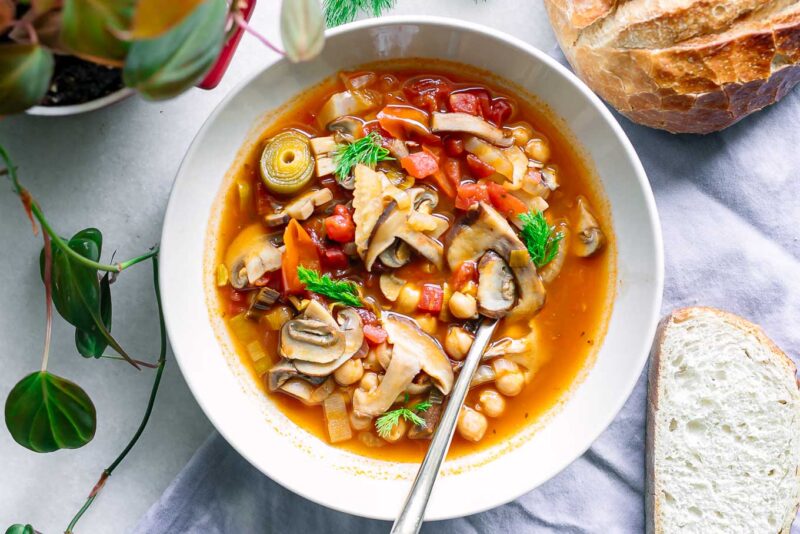 a white bowl of vegan cioppino soup with mushrooms on a table with sourdough bread for dipping