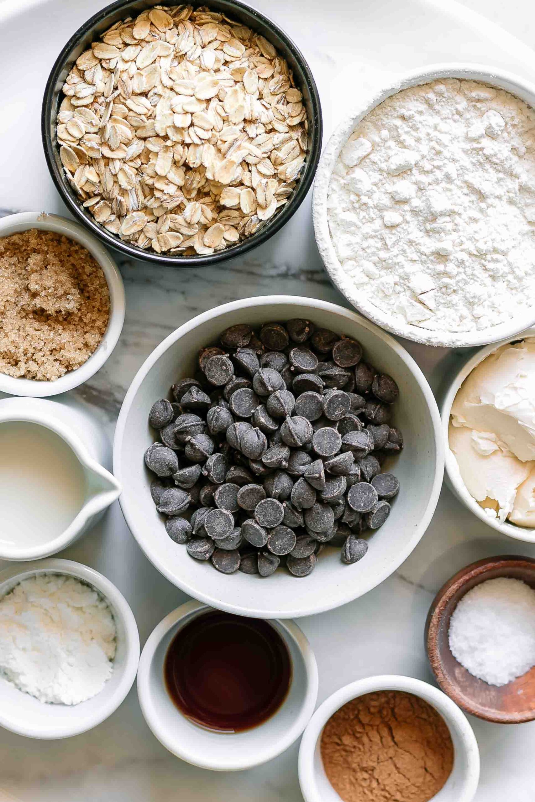 bowls of oats, flour, brown sugar, dark chocolate chips, and other ingredients for chocolate chip oatmeal cookies